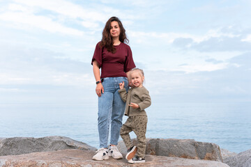 Obraz premium A young mother standing by the sea with her little daughter. The woman smiles gently as the child hugs her, with calm water and a cloudy sky in the background. Family, love, and lifestyle concept.