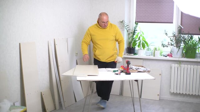 A man is focused on assembling furniture in a well-lit room. Surrounded by plants and tools, he carefully adjusts materials on the table, ensuring everything fits perfectly.