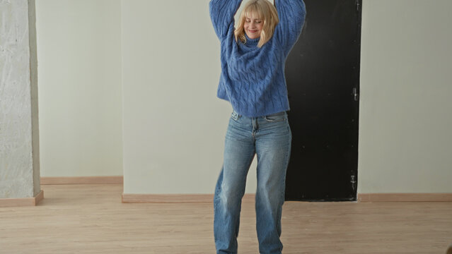 Woman in chunky blue sweater and jeans spins across an empty studio floor with arms raised and a bright smile, mid dance; joyful movement.
