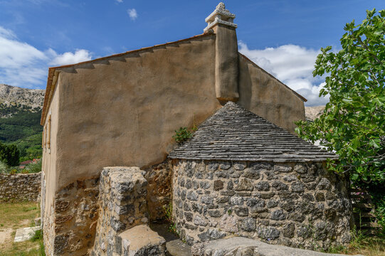Stone slab roof of a Romanesque church apse below a plastered gable with a carved finial, fig tree and blue sky, Adriatic island