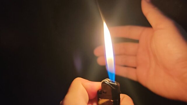 a portable lighter igniting in pitch black darkness. A bright yellow and blue flame burns steadily, casting a warm glow and illuminating a person's hand in the background. Concept of fire, survival