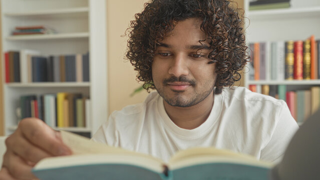 Man reads book and turns a page at a table in a library building with bookshelves behind him; concentration study.