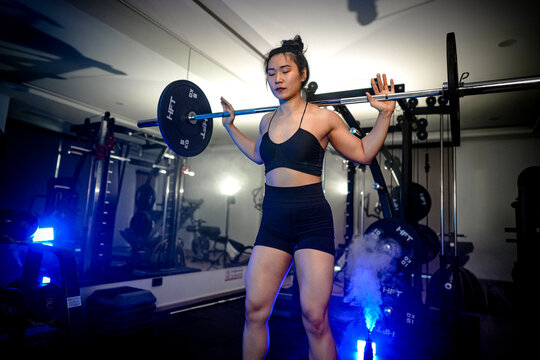 Women's strength and empowerment: an athletic Asian woman training her legs with a barbell squat in a dimly lit gym, channeling quiet determination, core braced and strong