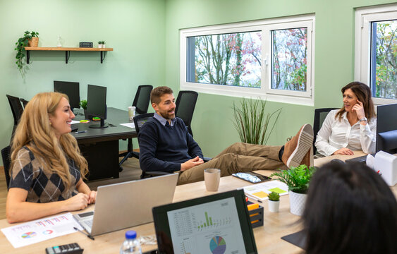 Seated man chats with smiling women while resting feet on their shared desk. Friendly office banter conveys humor, amusement and social connection during an informal break.