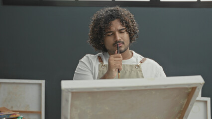 Man inspects paintbrush with raised hand while peering at canvas on easel in art studio  creative contemplation. © Krakenimages.com