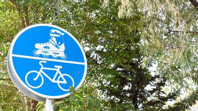 A round blue traffic sign indicating a combined path for roller skaters and bicyclists, positioned outdoors against a background of green tree branches