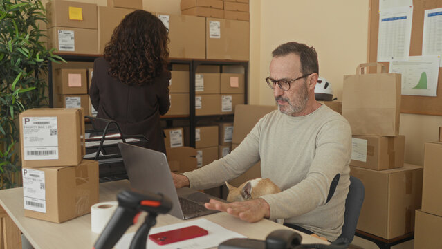 Man typing on laptop with small dog on lap and stacked shipping parcels, scanning labels with barcode reader in building; focused productivity.