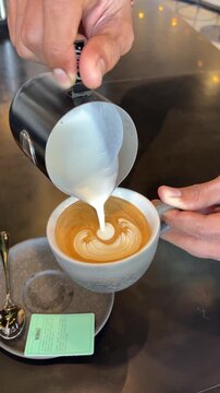 A hand pours steamed milk into a cup of espresso, creating a latte art design. The setting is a modern caf&eacute; with a sleek countertop.