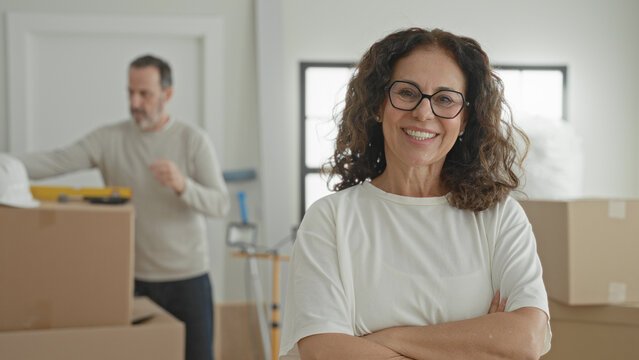 Middle aged woman smiling with arms crossed beside moving boxes while man handles helmet in building; contentment teamwork.