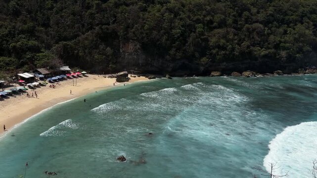 Aerial view of a sandy beach with gentle waves. Surrounded by lush greenery, beachgoers enjoy the sun. Small boats are visible in the water.
