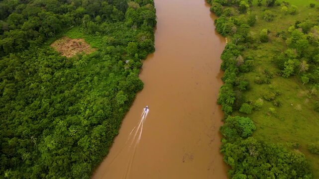 Panama, Darien. Aerial view shows a lone boat gliding down a muddy river. Lush green forests flank both sides of the winding waterway. The boat leaves a faint wake behind as it moves slowly forward