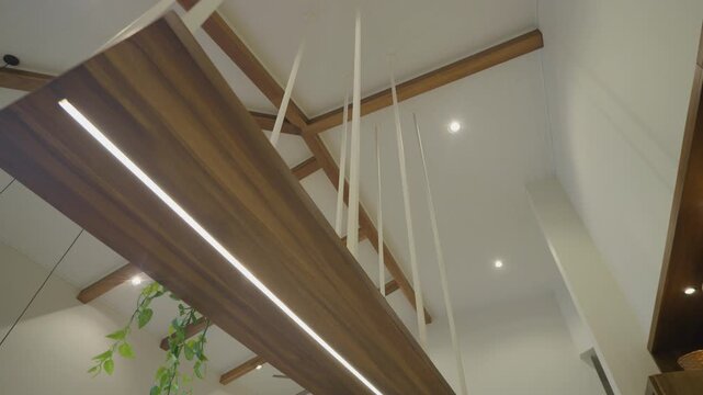 Modern room ceiling features white beams and a suspended wooden plank. Linear light strips run along the plank. Recessed ceiling lights illuminate the space. Green plant adds natural touch
