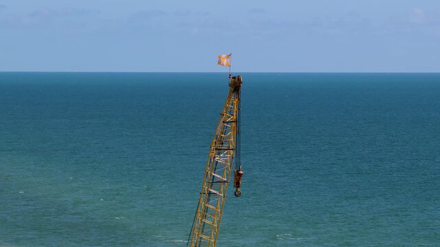 Side view of a tall metal construction crane boom with a hanging hook and checkered flag above a calm blue ocean.
