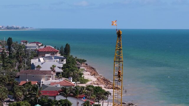 Elevated view of a tall yellow construction crane beside seaside houses along a turquoise coastline.