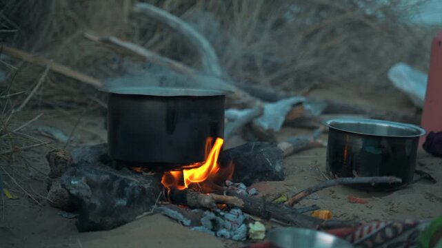Cinematic slow-motion static shot of a cooking pot boiling over an open wood campfire during outdoor desert camping in the Thar Desert near Jaisalmer, Rajasthan, India.