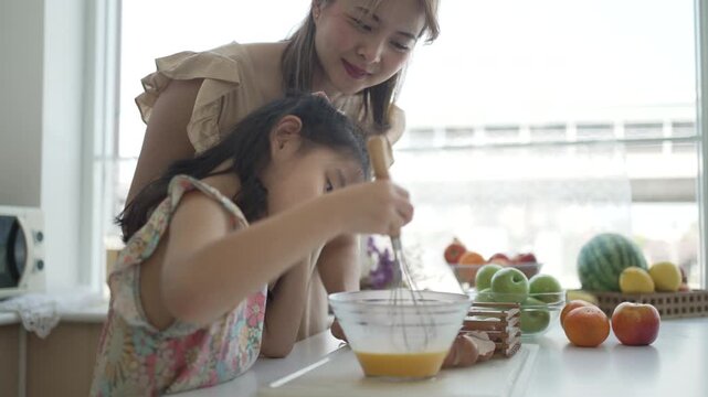 Mother laughing as daughter whisks egg mixture in bright kitchen