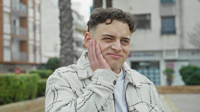 Man pressing cheek with hand in pained expression on a city street with apartment buildings; toothache discomfort.