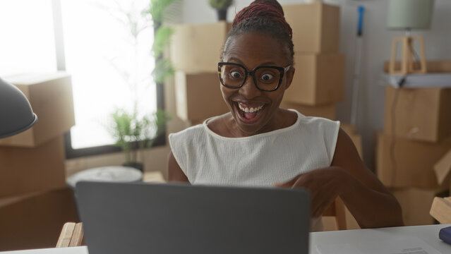 Young woman pumps fists and smiles widely at laptop surrounded by stacked moving boxes in a building; joy success.