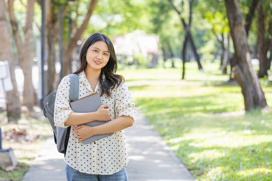 Young asian female university student carrying backpack and books smiling at campus park, happy college girl standing outdoors in nature with green trees and educational supplies