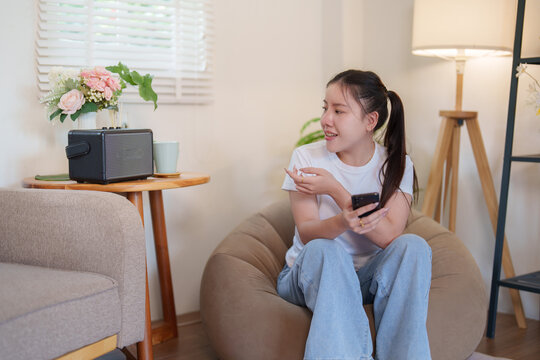 Asian woman controlling smart home speaker with smartphone application while relaxing in living room