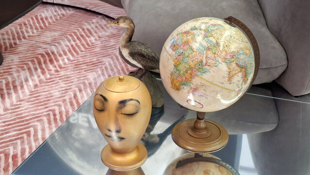 Vintage world globe map beside artistic golden face sculpture vase and taxidermy bird statue on reflective glass coffee table in modern luxury interior room on September 23, 2026 in Bangkok, Thailand.