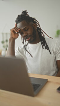 Young african american man with dreadlocks, hand to forehead over laptop at wooden desk in studio looking weary and focused; remote work fatigue.