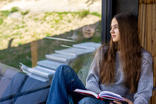 Young Woman Reading by Window With Reflection. Young woman sits by a large window holding an open book and gazing outside, with her reflection visible in the glass.