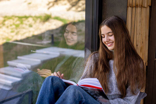 Smiling Woman Reading Book Beside Reflective Window. Smiling woman reads an open book beside a large window, with her reflection visible in the glass.