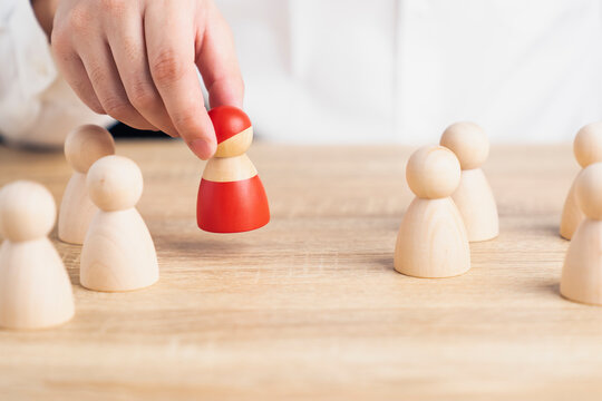 Talent recruitment and executive selection concept, businessman hand picking red wooden peg doll from group of natural figures, representing human resources hiring, staffing, and choice.