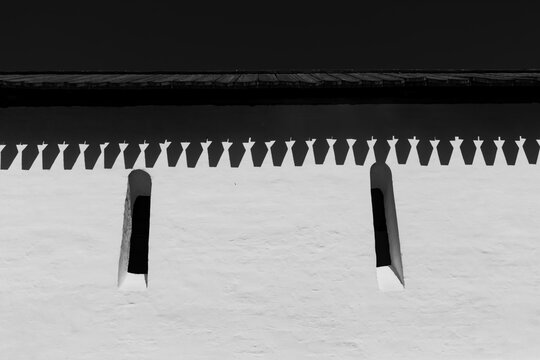 Old fortress monastery white defensive wall with narrow arrow slit windows and wooden shingle roof against a dark sky in a minimalist black and white architectural capture