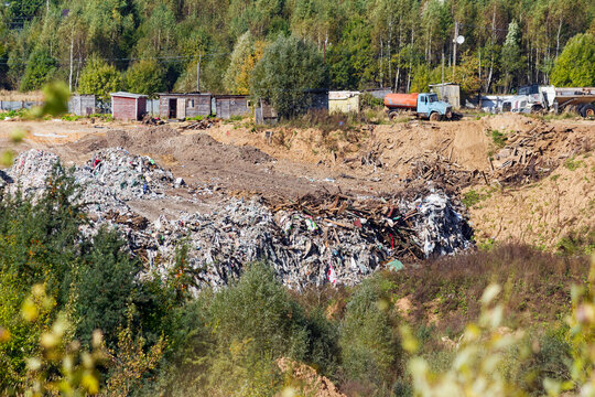 Large piles of trash and household waste scattered across an open rural landfill site with industrial trucks parked near old wooden sheds in the background