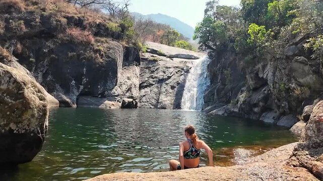 A woman wades into a natural pool and swims toward Likhubula Falls on Mount Mulanje, Malawi, as water cascades down the rocks ahead.
