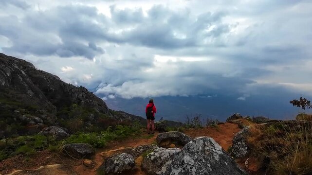 A woman with a backpack stops on a rocky overlook on Mount Mulanje, Malawi, and looks toward distant valleys beneath layered clouds.