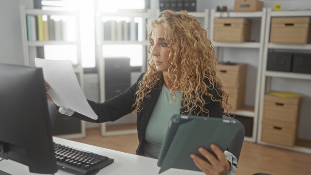 Woman middle aged blonde holding tablet and papers, hand visible, reading and comparing documents at office desk; calm concentration diligence.