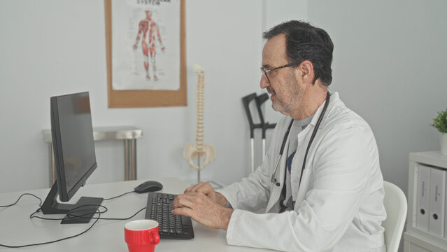 Man doctor typing on keyboard at computer desk with hands and stethoscope, wheelchair, spine model and red mug in clinic building; focused care.