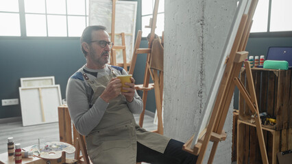 Man painter in apron sits at easel with palette, holding yellow coffee cup, hand on chin in studio  contemplative calm. © Krakenimages.com