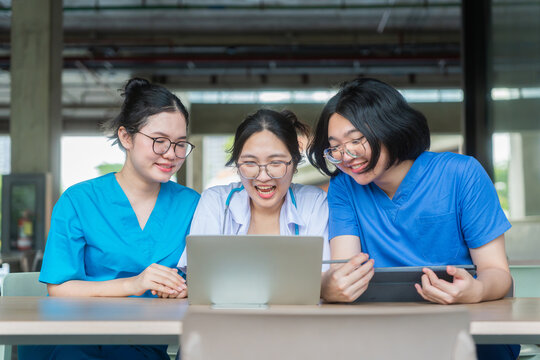 Medical students wearing scrubs and white coat using laptop and tablet together at university hospital, young doctors and nurses laughing and collaborating on project with digital devices in classroom