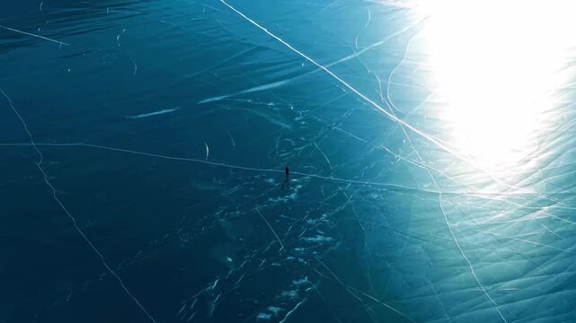 Aerial view of a person walking on deep blue cracked frozen lake ice with sun glare