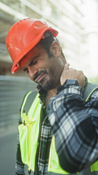 Man engineer in orange hardhat and reflective vest rubbing neck at a building site outdoors; workplace pain.
