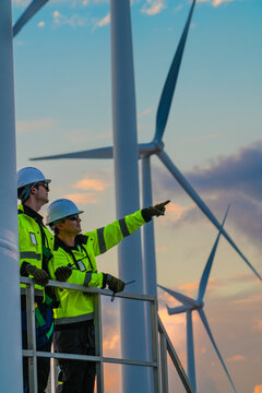 Two engineers on the turbine access platform watch a far machine spin from the walkway high on a wind turbine access platform in flaming sunset clouds at an operating renewable power.