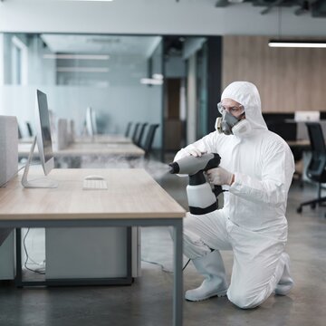 A worker wearing a full protective suit, mask, and goggles sprays disinfectant on an office desk with a computer, ensuring a sanitized workplace environment.