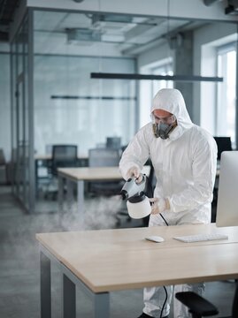 A worker wearing a full protective suit, mask, and goggles sprays disinfectant on an office desk with a computer, ensuring a sanitized workplace environment.