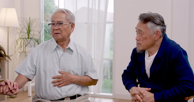 Senior Asian man gestures while discussing health and recovery needs beside another senior resident, receiving supportive nursing consultation inside modern assisted living community for wellness.