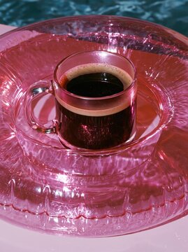 Black coffee in a glass mug resting on a pink inflatable pool float with water surface background