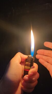 hands holding a portable lighter burning steadily in pitch black darkness. A bright yellow and blue flame illuminates the person's hands. Cinematic low light concept of fire, heat, survival, arson