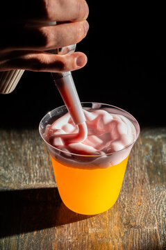 A professional bartender is using a whipping siphon to add pink foam topping onto a vibrant orange cocktail in a clear glass on a wooden table.