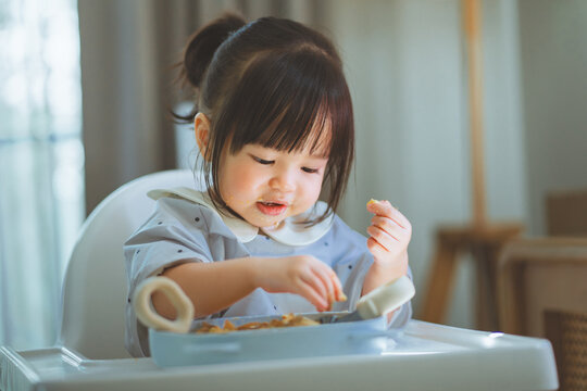 Toddler girl eating independently with hands while sitting in high chair at home, learning self feeding during mealtime, developing eating skills, independence, 