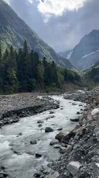 Rushing river flows through rocky valley floor. Mighty mountain peaks tower above verdant forested slopes. Clouds drift across sky, casting shadows over landscape
