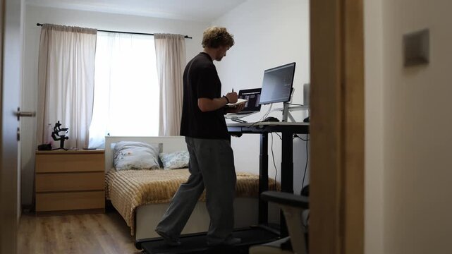 Young man reads a book while walking on a treadmill showing how staying active while working can be healthier than sitting at a computer desk for long hours every day