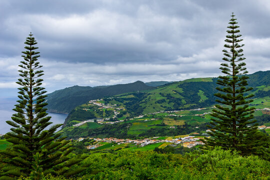 P&ocirc;r-do-Sol viewpoint on S&atilde;o Miguel island, showing two Norfolk Island pine trees framing a scenic coastal landscape with green hills, a small village, and the Atlantic Ocean under a cloudy sky